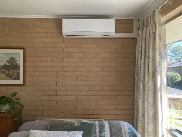 Wall-mounted split system above a bed on a brick bedroom wall with curtains and window in Greenwith